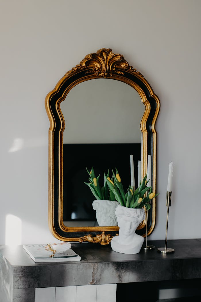 An ornate mirror with flowers, vase, and candles on a modern mantelpiece reflecting elegance.